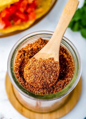 A wooden spoon scooping some taco seasoning from a glass jar.