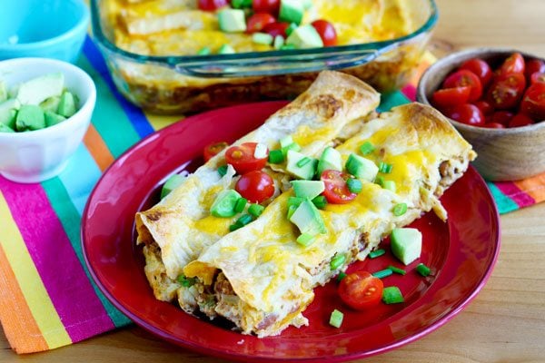 Enchiladas on a plate topped off with avocado, tomato, and green onion.