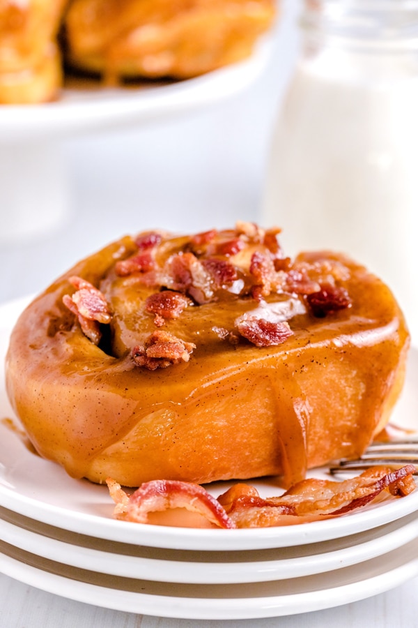 A close up picture of a sticky bun wit bacon with caramel glaze dripping down the size of the bun. 