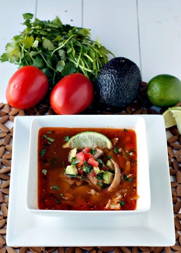 Chipotle Tortilla Soup in a white bowl surrounded by tomatoes, cilantro, avocados, and limes. 