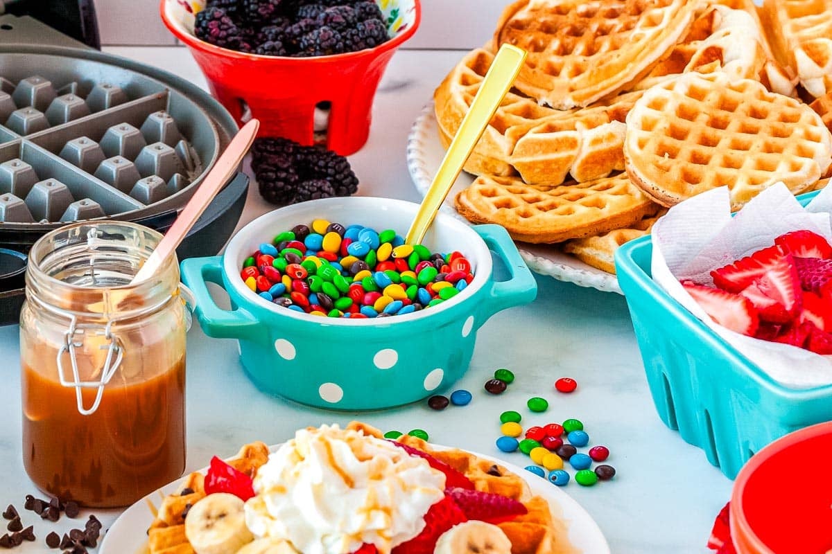 Waffles, a waffle iron, and toppings laid out on a counter.