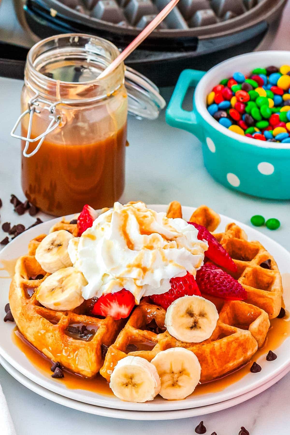 A Belgian Waffle on a white plate with tppings and a waffle iron in the background.