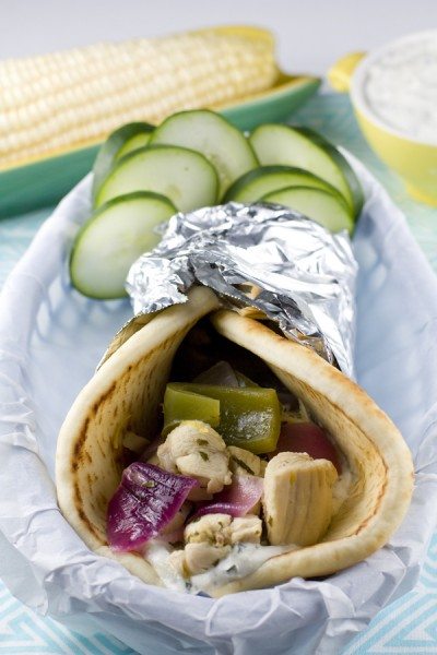Chicken Souvlaki in a pita with cucumbers and corn in the background.