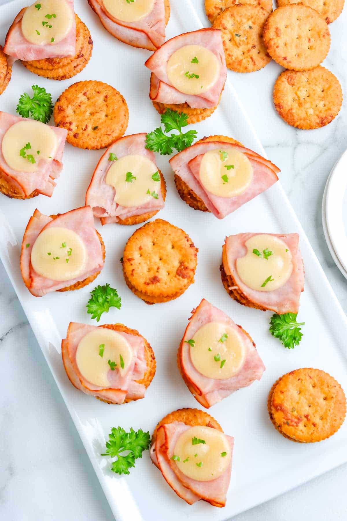 Ranch crackers on a white platter, some with ham and cheese on them. 