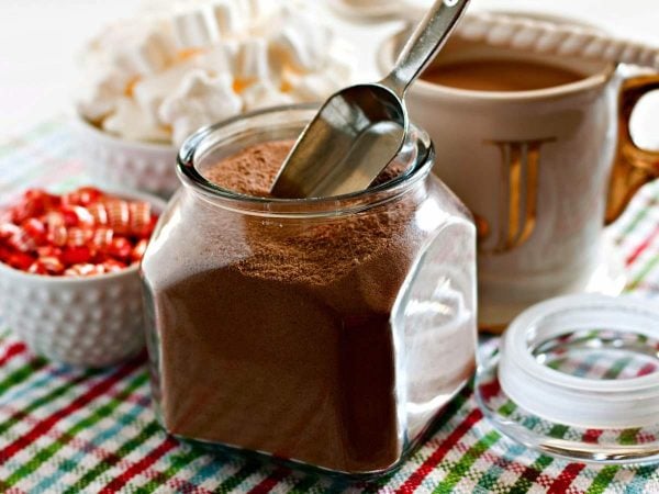 Hot chocolate mix in a glass jar with a cup of hot chocolate and a bowl of marshmallows in the background. 