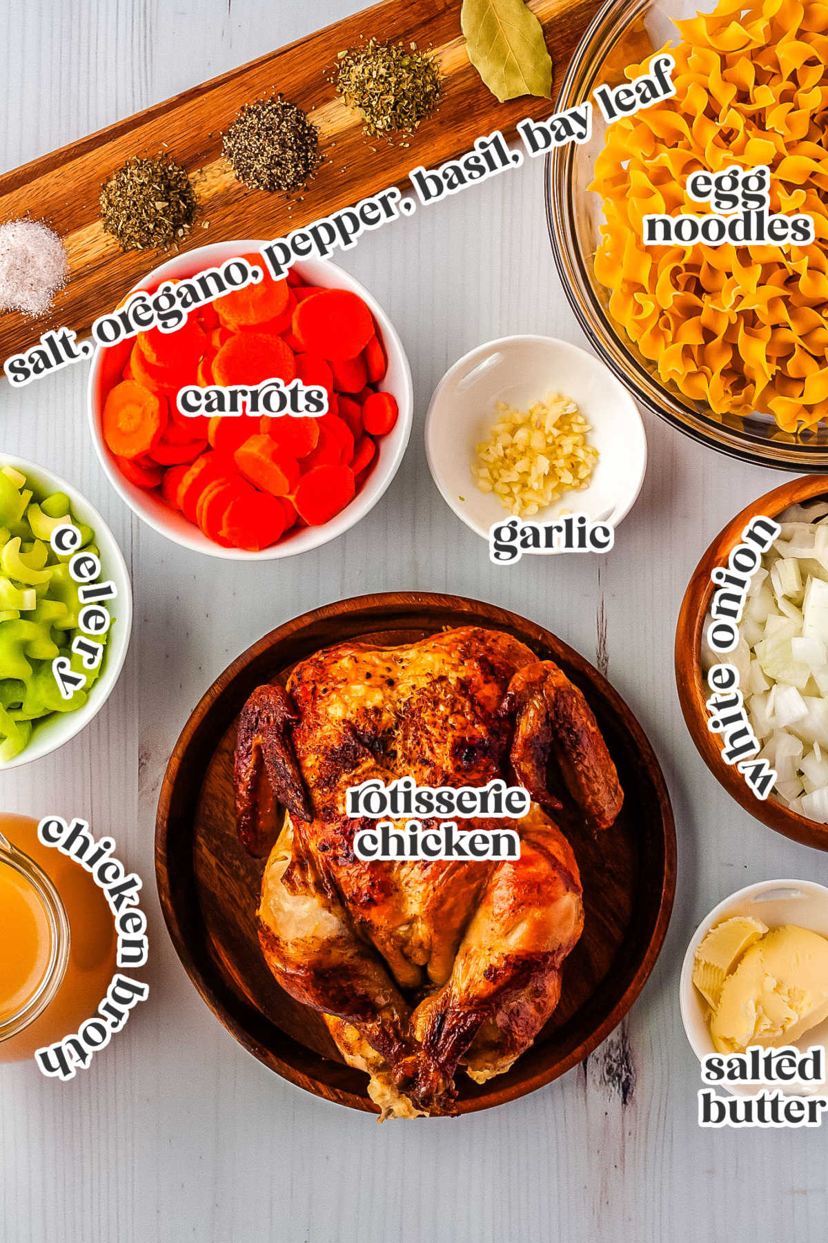 A flat lay of ingredients needed to make rotisserie chicken noodle soup including a whole rotisserie chicken, chopped celery, sliced carrots, minced garlic, egg noodles, chopped white onion, butter, chicken broth, and spices.