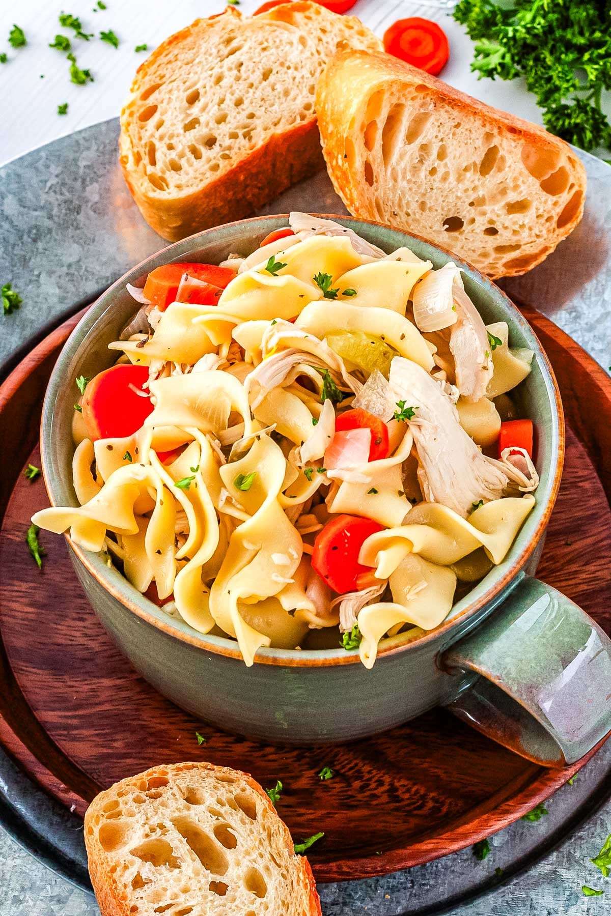 A green bowl filled with chicken noodle soup using rotisserie chicken resting on a wooden plate next to two slices of crusty bread.