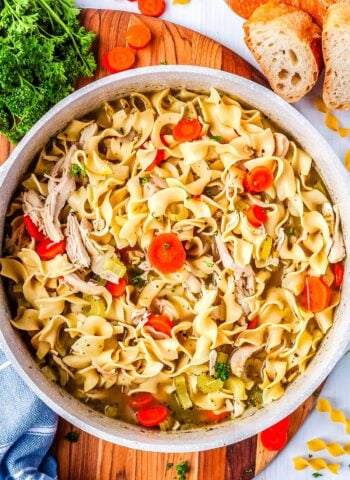 An overhead view of a large pot filled with chicken noodle soup with rotisserie chicken surrounded by fresh parsley and slices of bread.