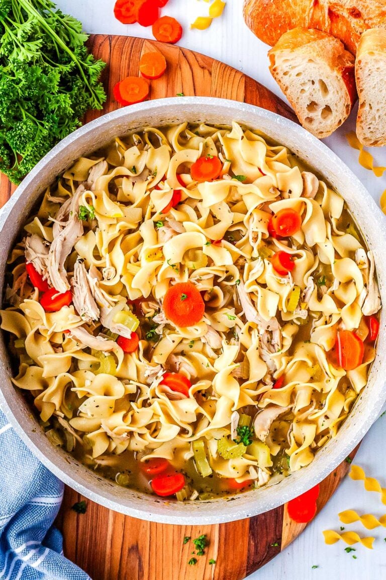 An overhead view of a large pot filled with chicken noodle soup with rotisserie chicken surrounded by fresh parsley and slices of bread.