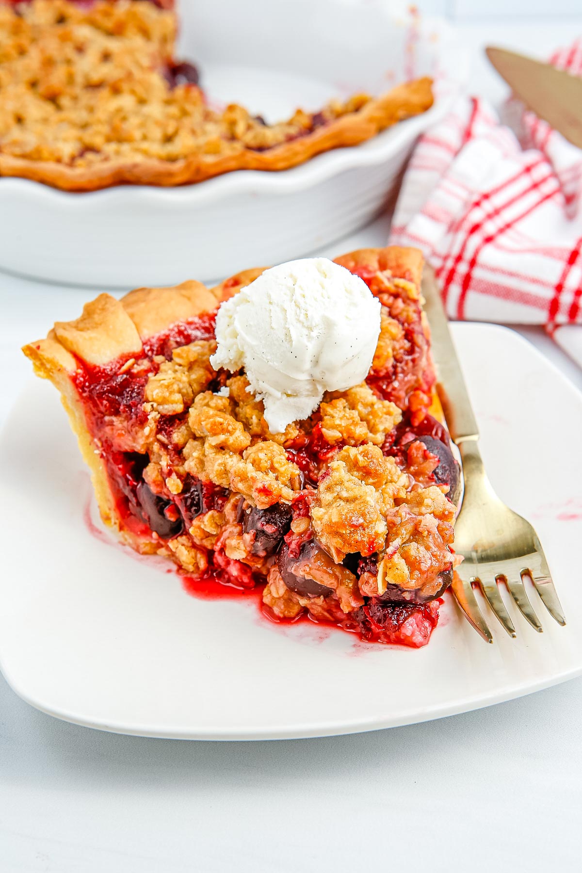 A sliced of the finished Cherry Crumble Pie on a white plate with a scoop of ice cream on top.