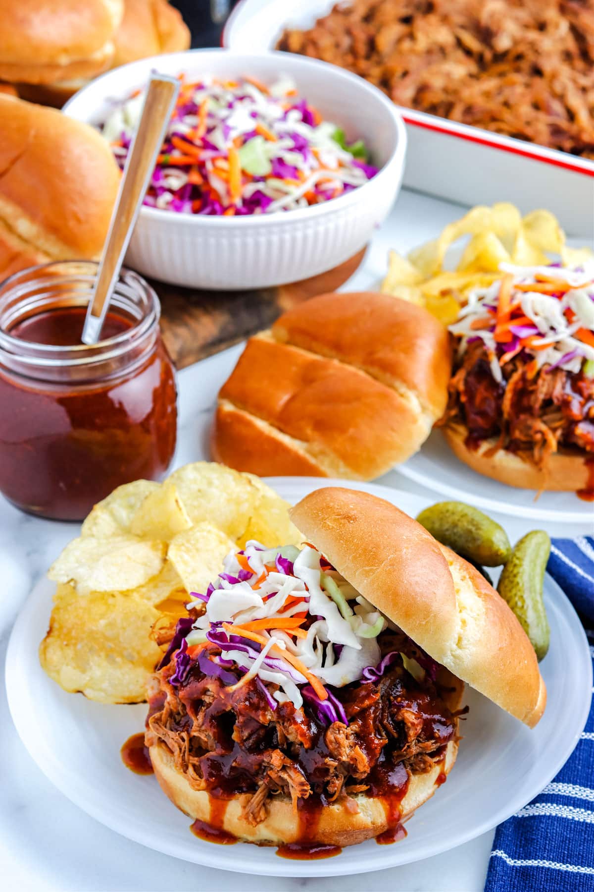 A picture of the Pulled Pork on a roll on a plate with potato chip and pickles on the side of the plate.