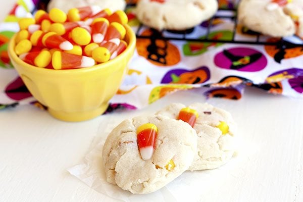Two sweet and salty candy corn cookies with a bowl of candy corn and more cookies on a wire rack in the background