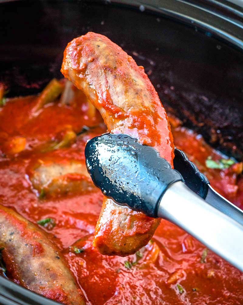Tongs picking up an Italian sausage cooked in a slow cooker with onions, peppers, and sauce. 