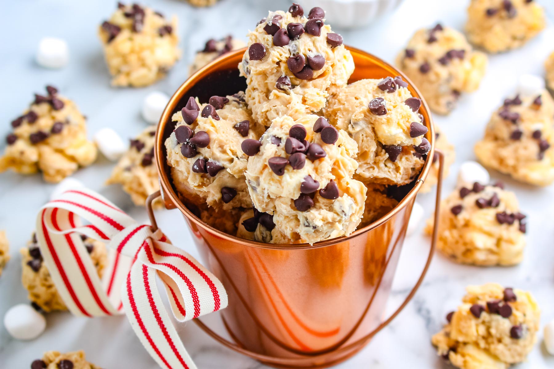 A copper bucket filled with no bake avalanche cookies topped with mini chocolate chips with more cookies scattered on a marble surface.