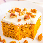Slice of pumpkin snacking cake topped with cream cheese frosting and candied pecans on a white plate, with crumbs scattered around.