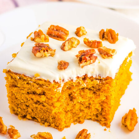 Slice of pumpkin snacking cake topped with cream cheese frosting and candied pecans on a white plate, with crumbs scattered around.
