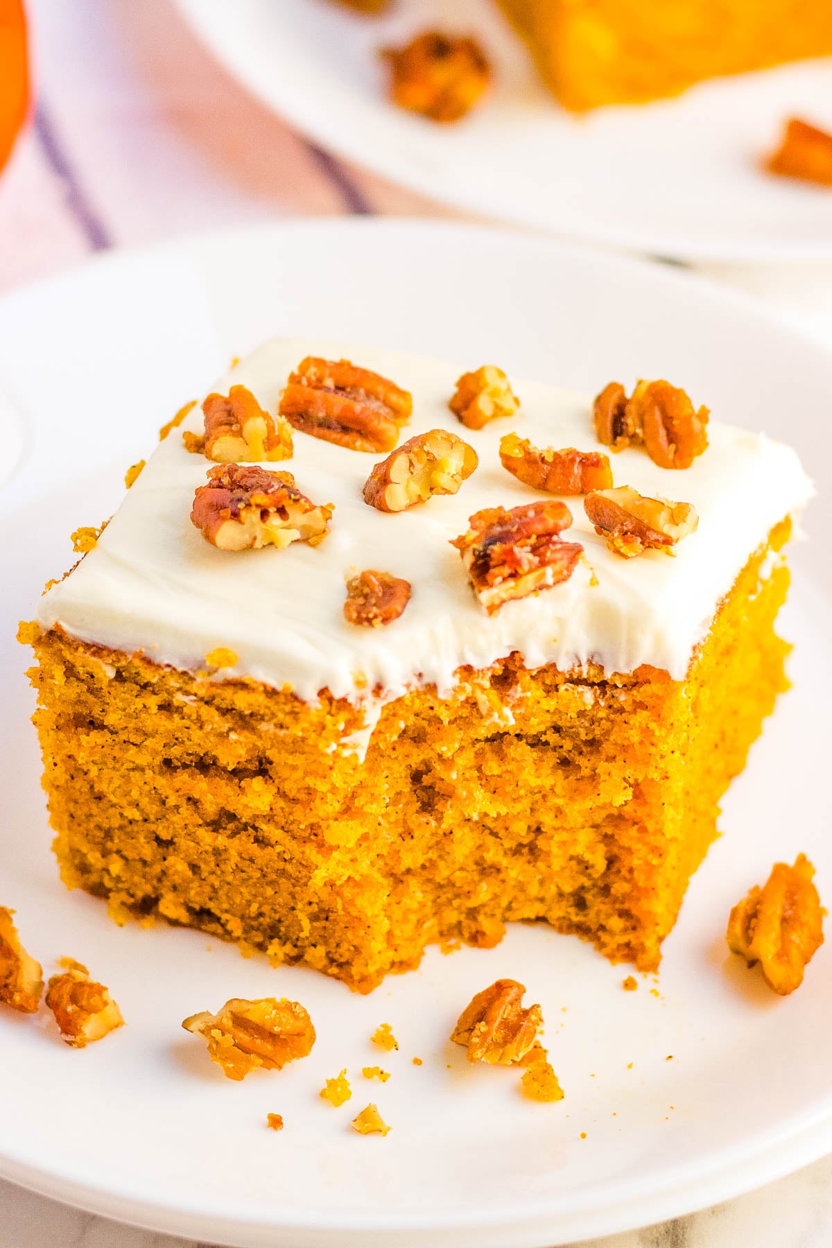 Slice of pumpkin snacking cake topped with cream cheese frosting and candied pecans on a white plate, with crumbs scattered around.