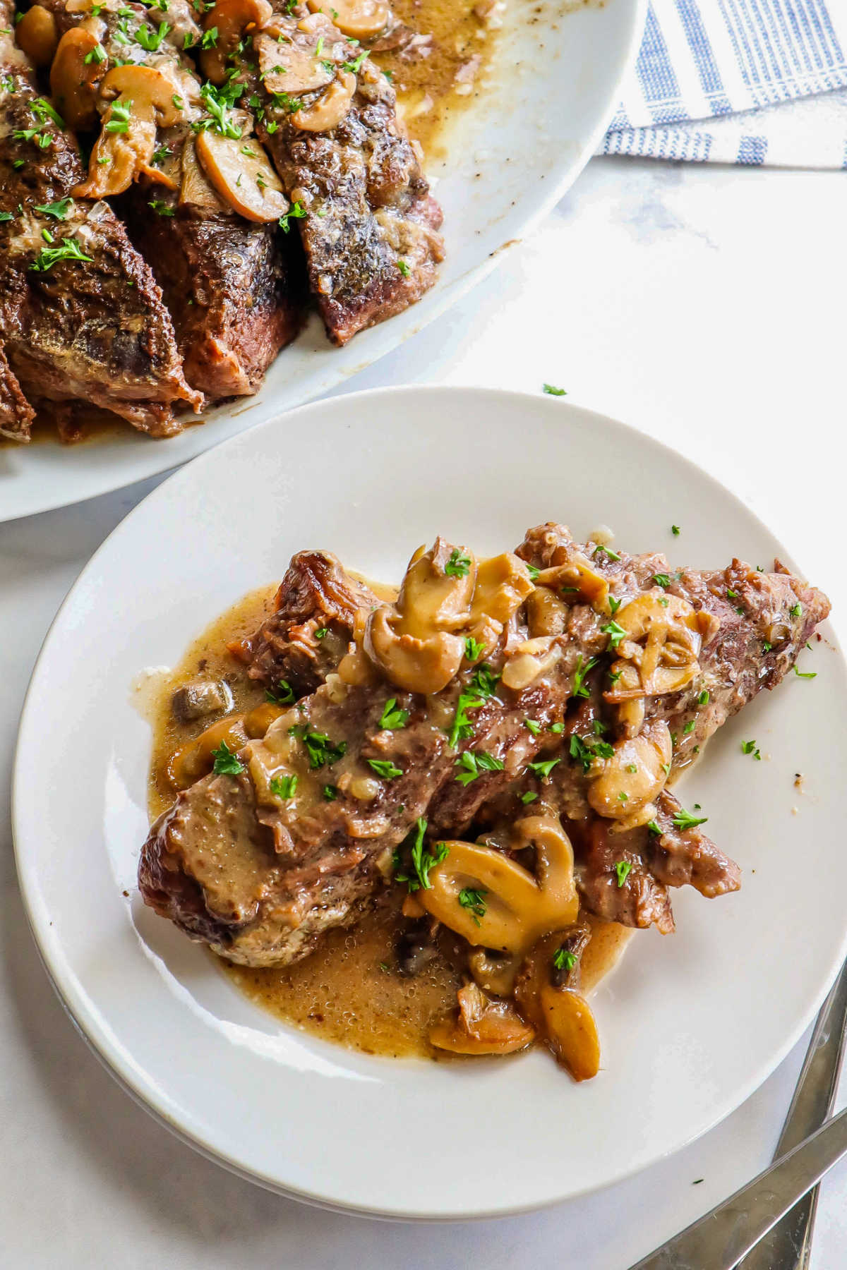 An overhead picture of pot roast with mushrooms on a white plate and a platter of pot roast in the background.