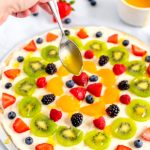 A spoon drizzling a glaze over the fruit pizza.