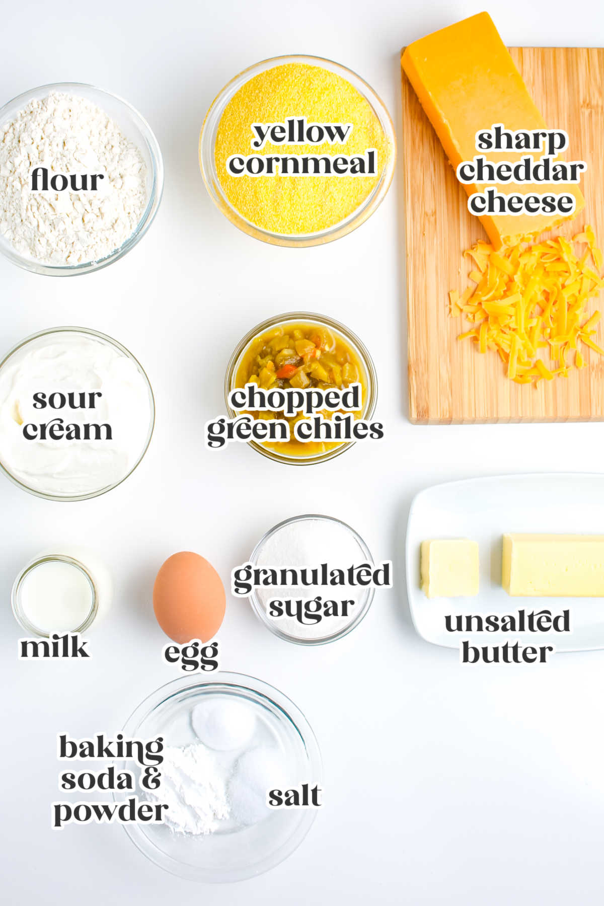 Ingredients for cheesy green chile cornbread including cornmeal, flour, cheddar cheese, sour cream, diced green chiles, butter, egg, and milk.