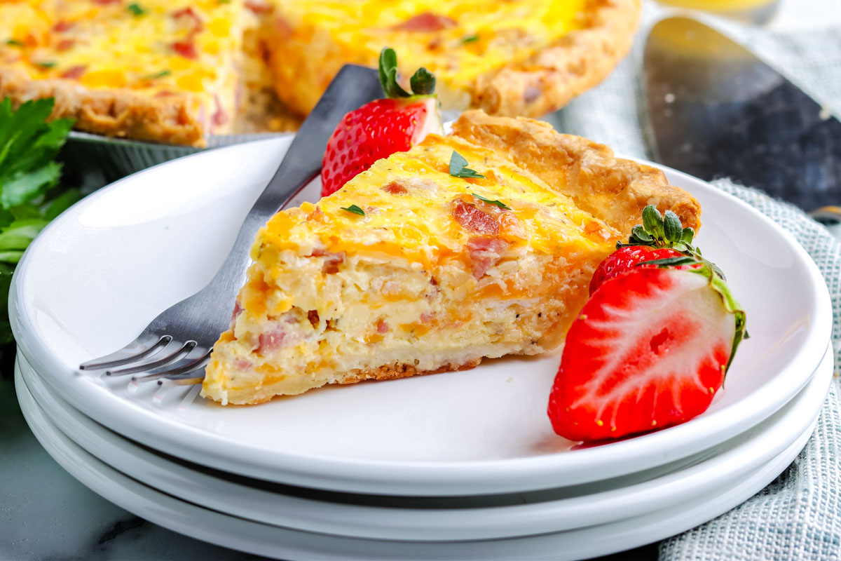 A slice of ham and cheese quiche on a white plate with strawberries on the plate, too.