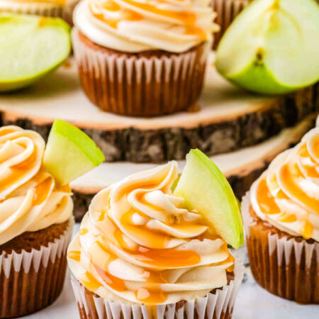 Caramel apple cupcakes on a wooden board, topped with caramel frosting, drizzle, and apple slices for garnish.