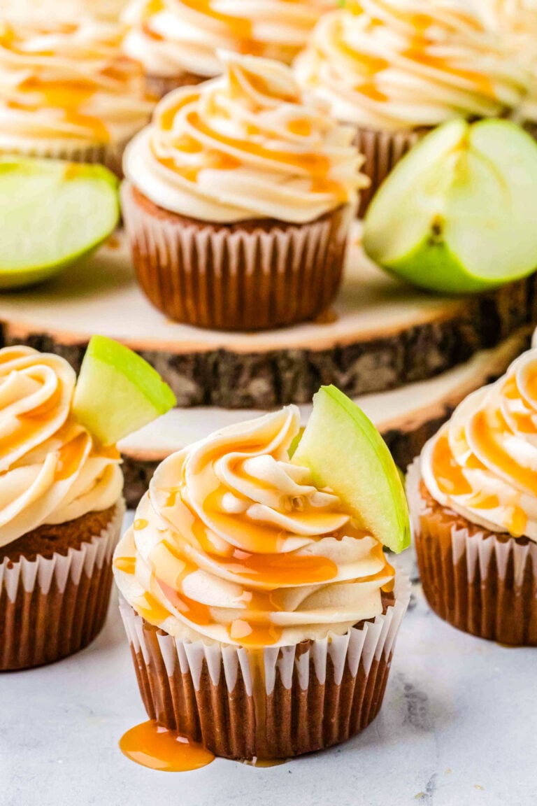 Caramel apple cupcakes on a wooden board, topped with caramel frosting, drizzle, and apple slices for garnish.