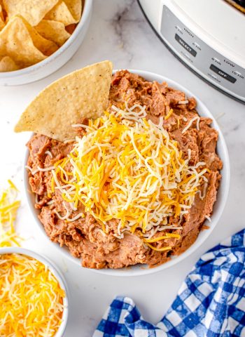 An overhead picture of the finished Mexican Refried Beans in a white bowl that's garnished with shredded Mexican cheese and tortilla chips.