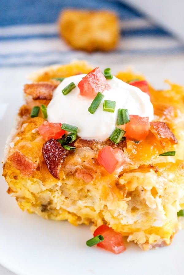 A piece of tater tot casserole on a white plate that's topped with sour cream, tomatoes, and chives.