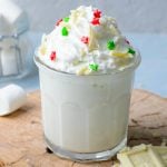 White hot chocolate in a clear glass that's garnished with whipped cream, white chocolate, and sprinkles.