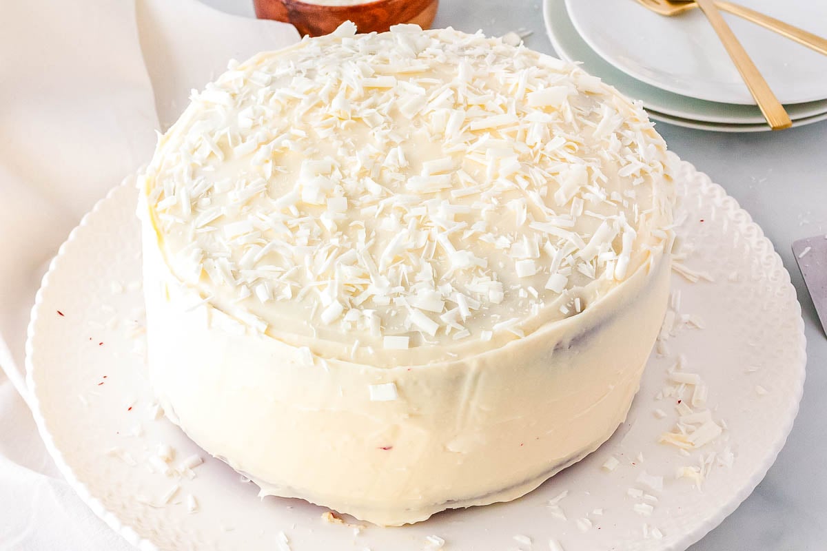 A whole Red Velvet Cheesecake Cake on a white platter, generously covered in cream cheese frosting and garnished with white chocolate shavings.