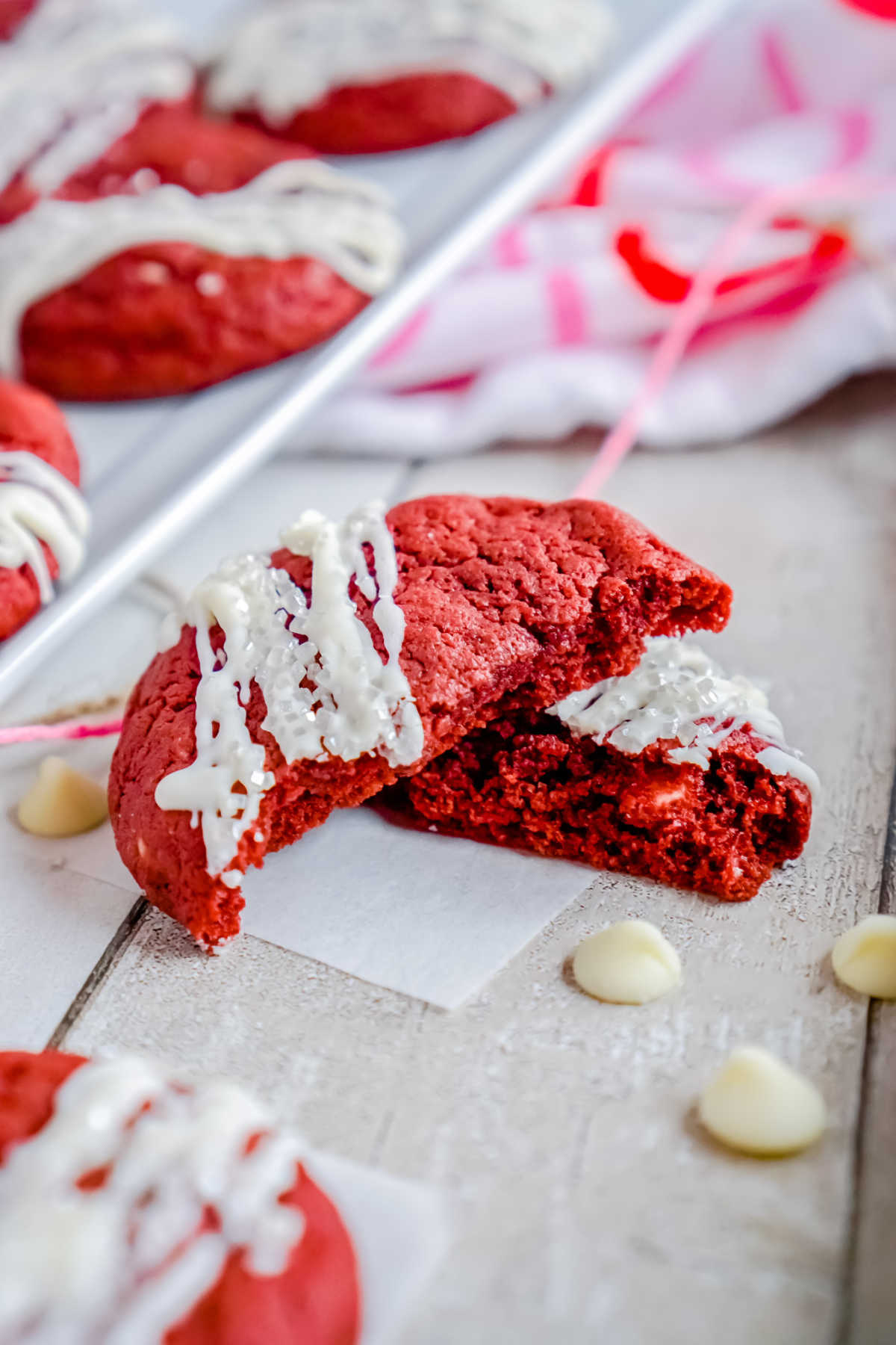 A chewy Red Velvet Cookie broken in half sitting on a table with white chocolate chips all around.