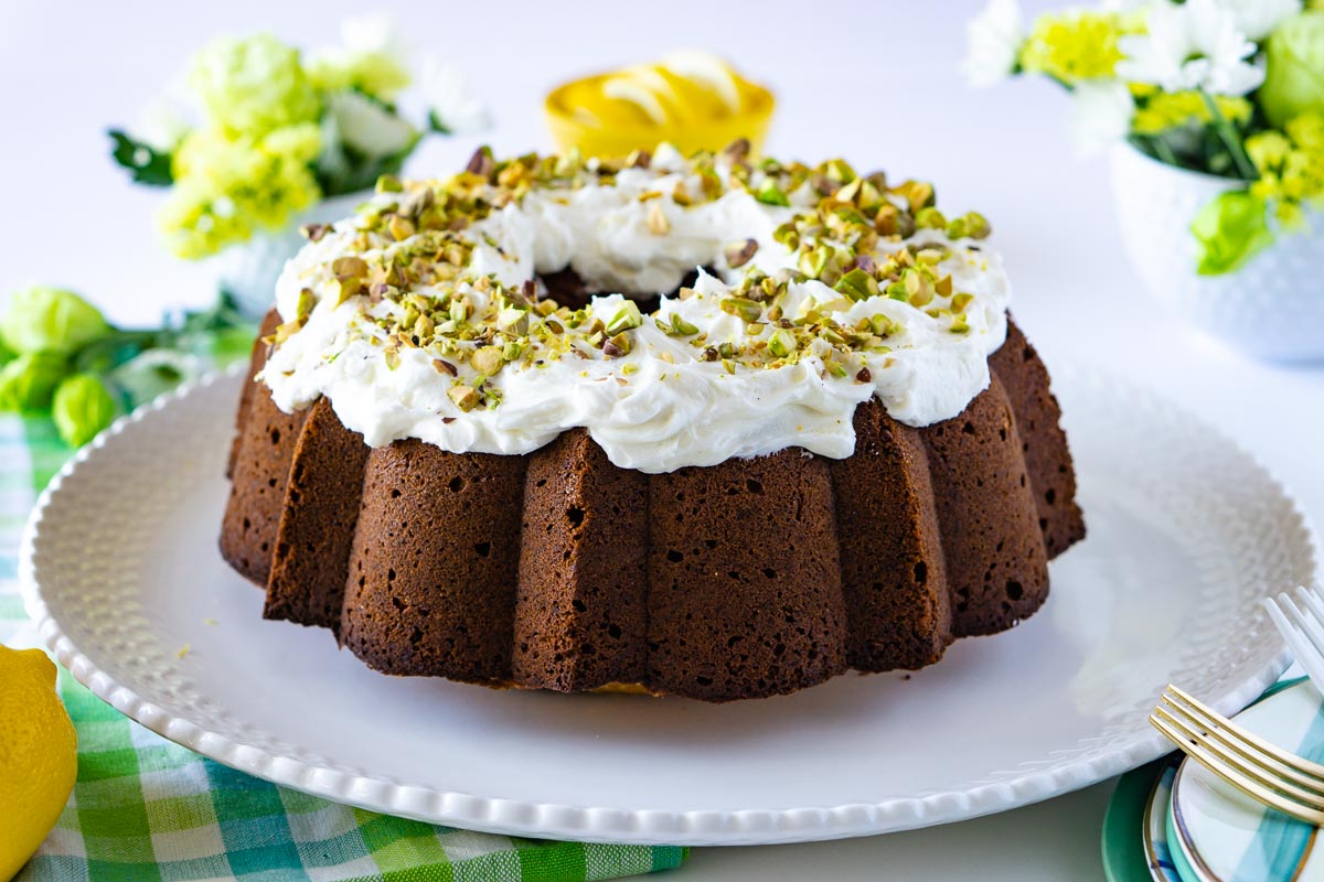 The entire Pistachio Bundt Cake on a white cake plate. It has lemon frosting on top and chopped pistachios .