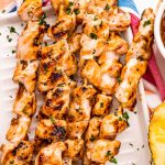 Chicken skewers on a white platter with pineapple teriyaki sauce nearby.