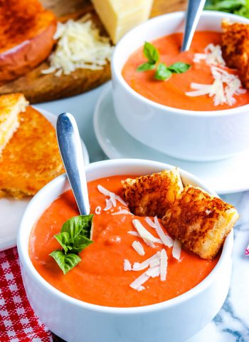 Tomato Soup in white bowls garnished with small grilled cheese cubes and basil.