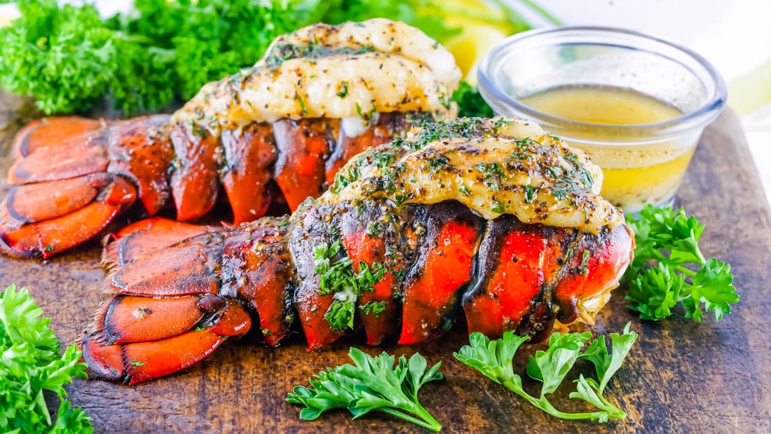 A close up picture of two Smoked Lobster Tails on a cutting board with a dish of melted butter for dipping the lobster meat into.