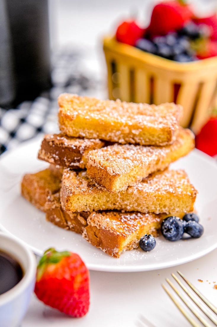 A pile of Air Fryer French Toast Sticks on a white plate. The French Toast sticks have been dusted with powdered sugar.