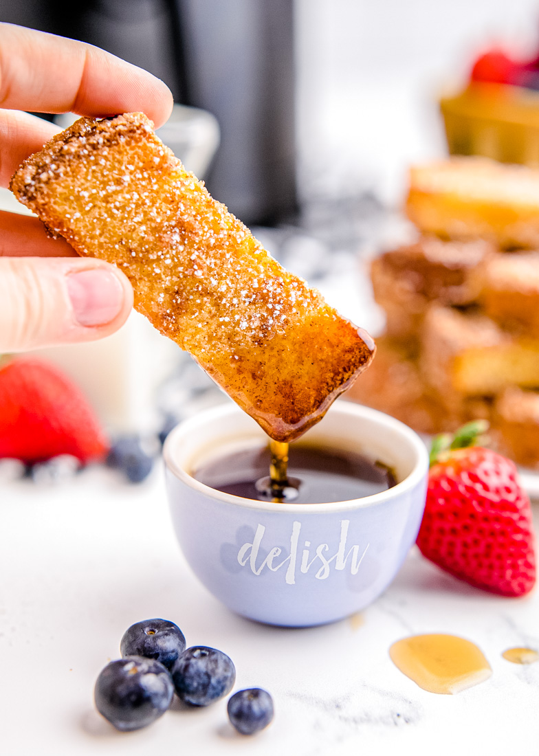 A French toast stick that's been cooked in the air fryer being dunked in syrup.