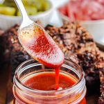 A spoon drizzling some of the Texas BBQ Sauce from a glass jar.