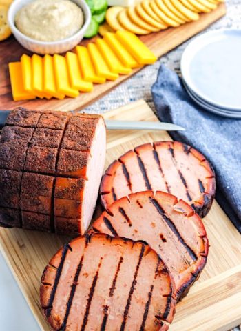 Three slices of smoked bologna beside their whole counterpart on a wooden board.