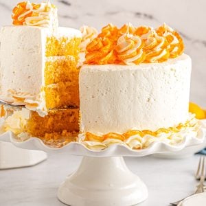 The finished Orange Creamsicle Cake recipe on a white cake stand with a slice being taken out of it.