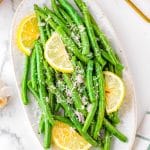 The finished Air Fryer Green Beans on a white platter garnished with Parmesan and lemon wedges.