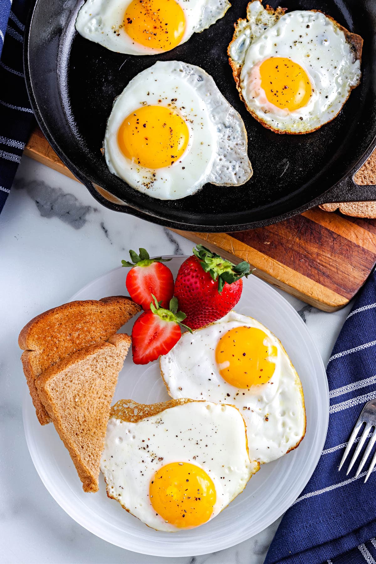 An overhead picture of fried eggs on a white plate and in an iron skillet. 