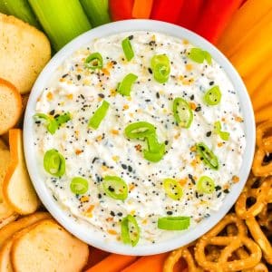 A close up picture of the Everything Bagel Dip in a white serving bowl surrounded by bagel chips and veggies.