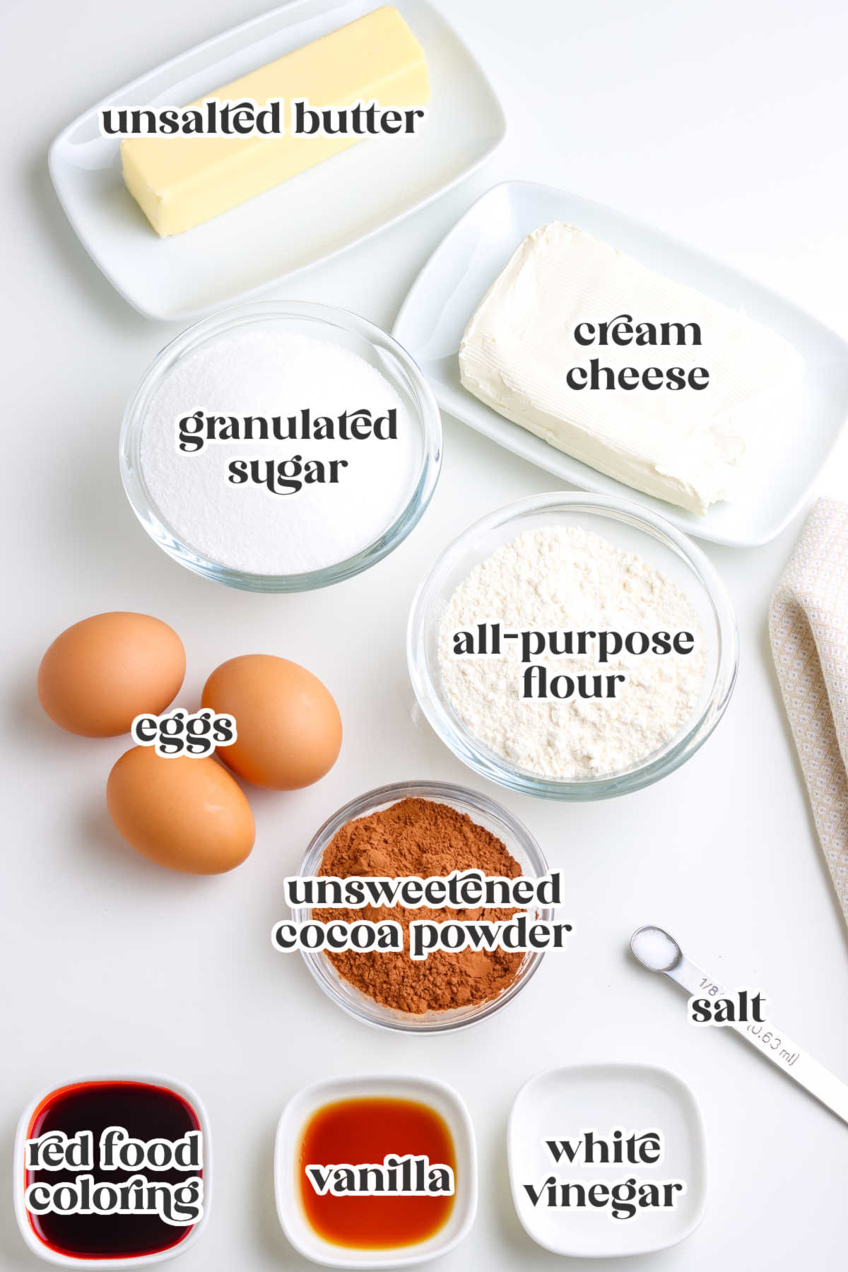 Overhead view of butter, sugar, cream cheese, eggs, flour, cocoa powder, red food coloring, vanilla, vinegar, and salt neatly arranged on a white background.