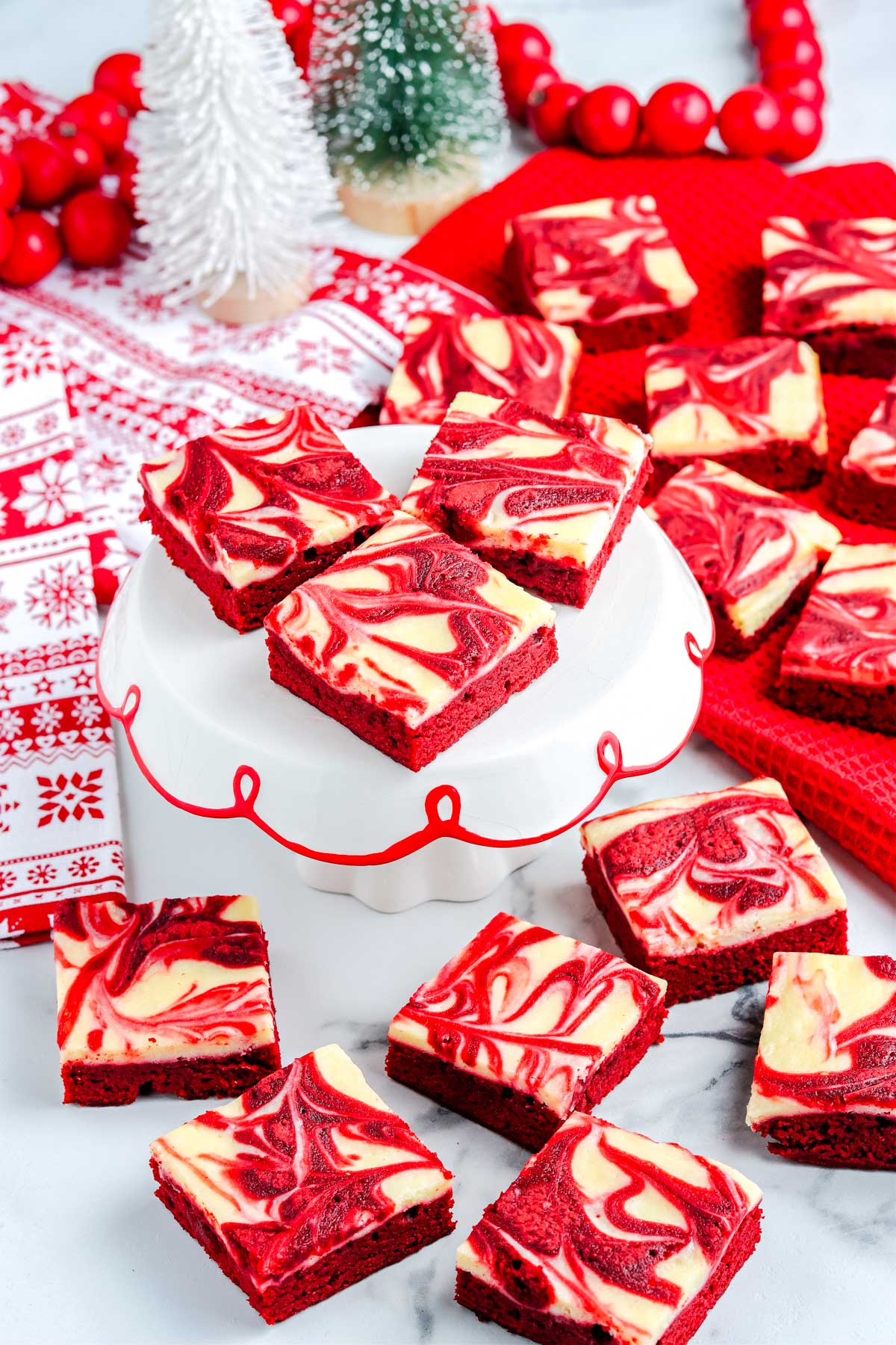 Red velvet brownies displayed on a white cake stand with festive decorations including snowy bottle brush trees, red garlands, and seasonal linens.