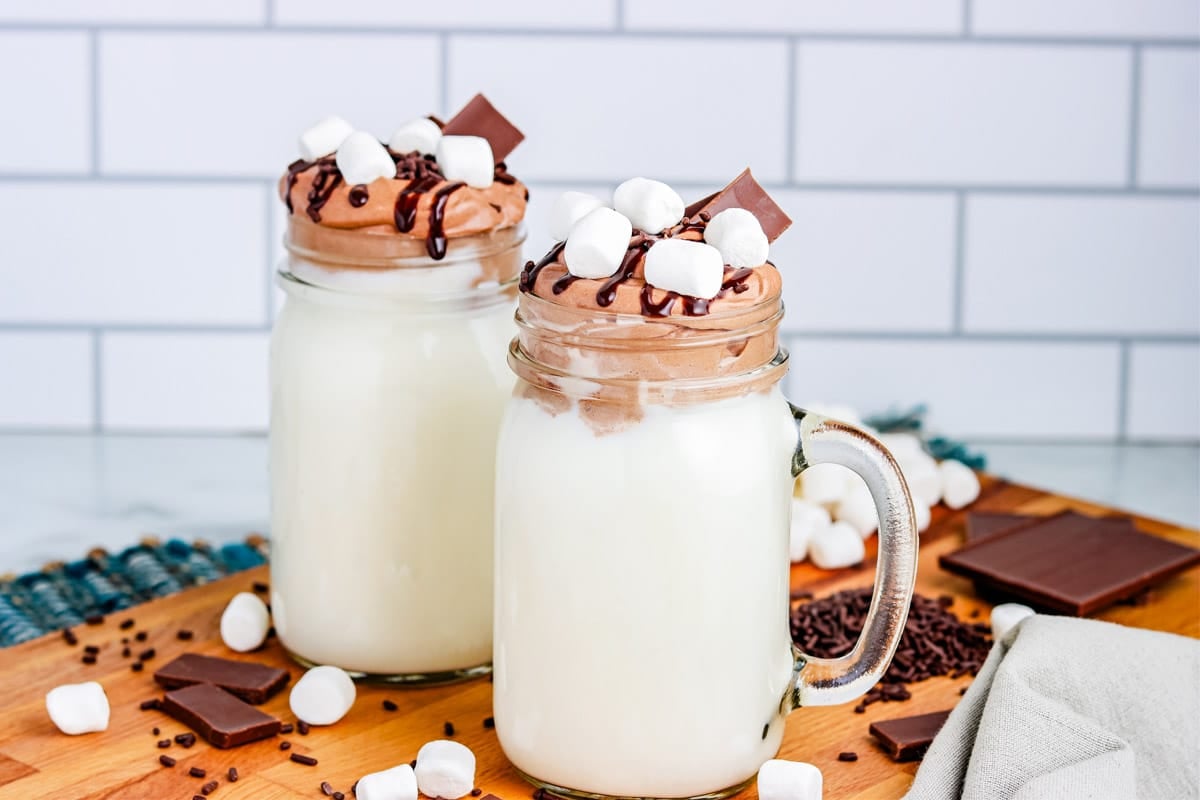 Two mugs of Whipped Hot Chocolate surrounded by marshmallows and pieces of chocolate.