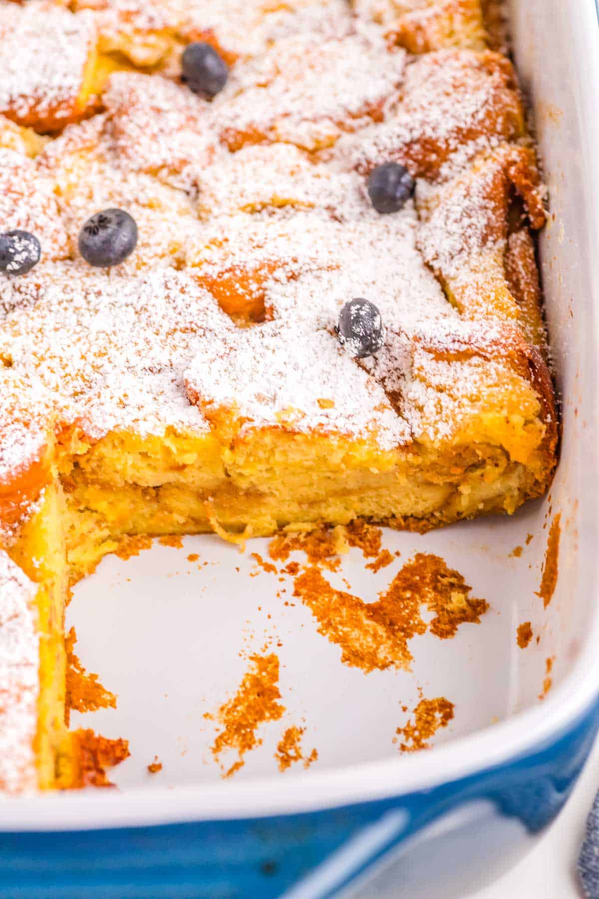 Brioche French Toast Casserole in a blue casserole dish garnished with powdered sugar and blueberries.
