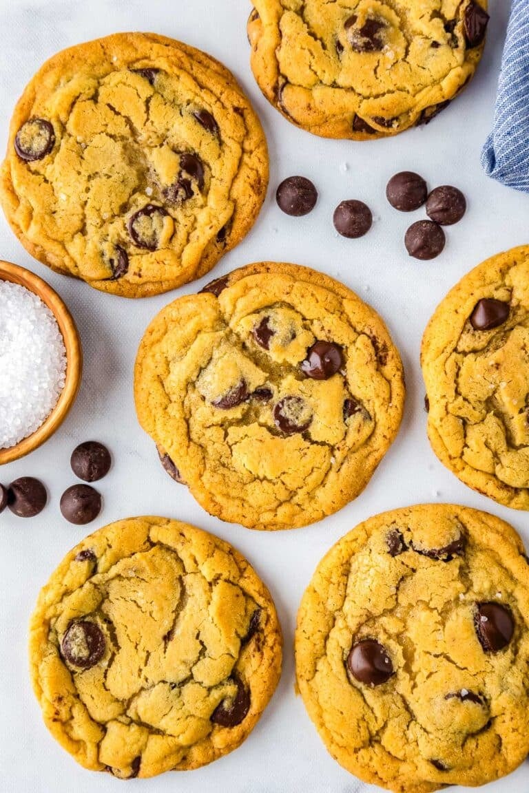 Six bread flour chocolate chip cookies on a counter with chocolate chips sprinkled around them.
