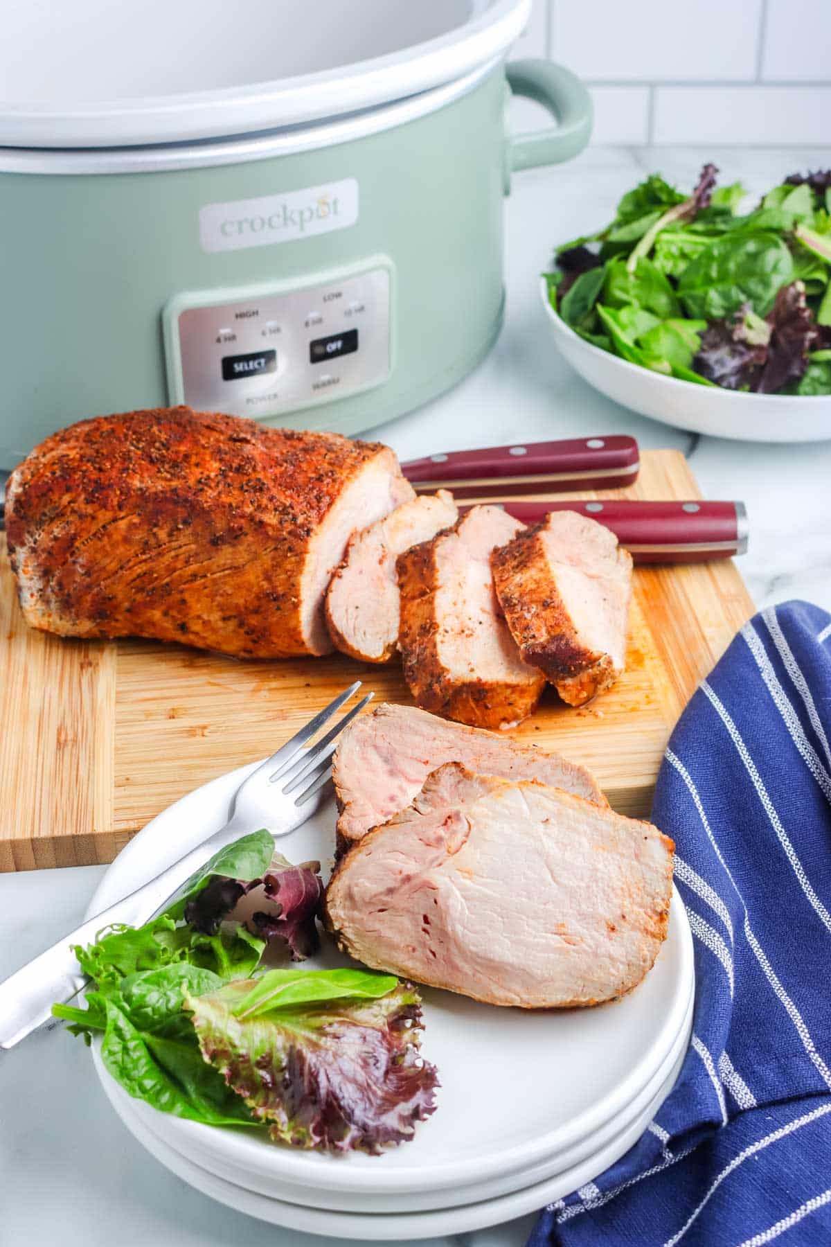 Sliced crockpot pork loin in the background with slices on a white plate.